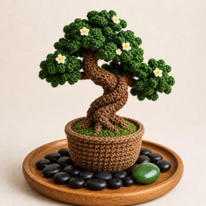 Könnte beinhalten: Ein handgehäkelter Bonsai-Baum mit einem gedrehten braunen Stamm, grünen Blättern und kleinen weißen Blüten. Der Baum steht in einem braunen Topf auf einem Holztablett mit schwarzen Steinen und einem grünen Stein.