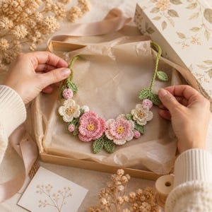 Può includere: Una collana fatta a mano all'uncinetto con fiori rosa e crema, centri gialli e foglie verdi. La collana floreale è presentata in una confezione regalo con un design floreale. La collana è fatta di filato.