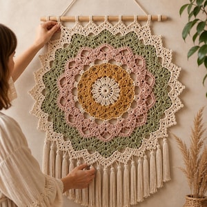 Puede incluir: Un tapiz de pared redondo de ganchillo hecho a mano con un diseño de mandala. La pieza presenta círculos concéntricos de color crema, verde, rosa y dorado. Está suspendido de una barra de madera con una borla de color crema en la parte inferior. Aproximadamente 76 cm de diámetro.