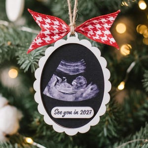 May include: An oval-shaped Christmas ornament with a scalloped white frame, featuring a black center with a fetal ultrasound image and the text "See you in 2027" printed in white. A red and white houndstooth ribbon bow is tied at the top.