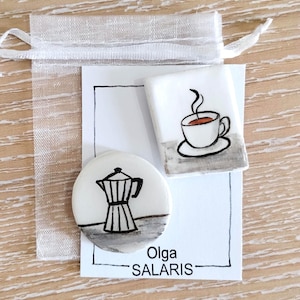May include: Two white ceramic pins featuring black ink illustrations of a moka pot and a coffee cup with steam. The pins are attached to a white card with the text Olga SALARIS, presented inside a sheer white organza drawstring gift bag.