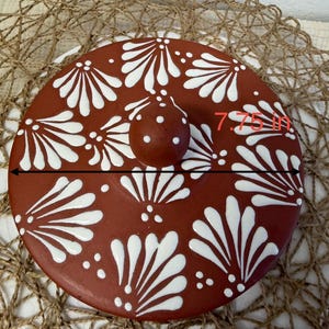 May include: A round, terracotta-colored ceramic lid featuring a white hand-painted floral pattern and a central knob handle. The lid measures 7.75 inches in diameter, as indicated by the text and measurement line visible on the surface.