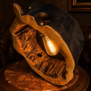 Op de afbeelding: Een tafellamp gemaakt van een dik, gebogen stuk donker, verkoold hout met een natuurlijke, ruw getextureerde binnenkant. Een Edison-stijl gloeilamp hangt aan de bovenste binnenbocht en verspreidt een warme, gouden gloed over de houtnerf.