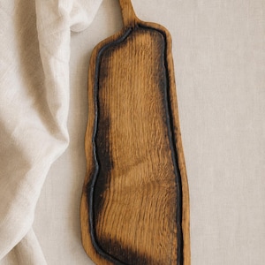 Può includere: Un tagliere da portata in legno lungo e stretto con un bordo naturale irregolare e un manico prominente sulla parte superiore. Il tagliere presenta un bordo scuro con effetto bruciato che circonda una superficie centrale con venature chiare.