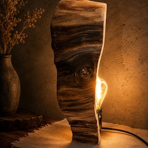May include: A table lamp featuring a vertical slab of natural dark brown wood with a prominent knot. The lamp includes a single exposed Edison-style light bulb emitting a warm glow, a brass-colored socket, and a black power cord extending outward.
