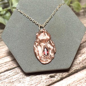 May include: A necklace featuring a metallic copper-toned pendant with an irregular, textured surface and a single marquise-cut deep red gemstone set in the center. The pendant hangs from a simple silver-toned metal chain link necklace.