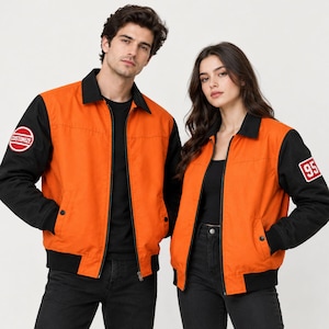 Könnte beinhalten: Zwei Jacken mit leuchtend orangefarbenem Rumpf und schwarzen Ärmeln, Kragen und Saum. Die linke Jacke hat einen runden Aufnäher mit dem Text CUSTOMIZE auf dem Ärmel, während die rechte Jacke einen rechteckigen Aufnäher mit der Zahl 95 aufweist.