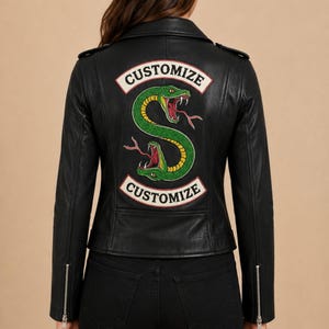 Op de afbeelding: Een zwart leren jack met een grote geborduurde patch op de achterkant. De patch toont een groene slang in een S-vorm met rode tongen en hoektanden, geflankeerd door twee witte banieren met de zwarte tekst CUSTOMIZE.