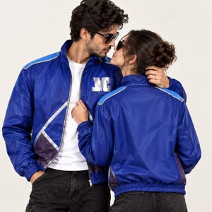 Puede incluir: Una chaqueta azul real con cremallera, ribetes reflectantes plateados, cuello alto y las letras MC en plata en el pecho izquierdo. La chaqueta tiene mangas largas con puños elásticos y dobladillo, combinada con una camiseta blanca y jeans grises.