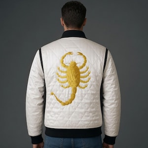 May include: A white quilted bomber jacket featuring a large, embroidered golden yellow scorpion on the back. The jacket has a black ribbed collar, black ribbed cuffs, and a black ribbed hem, worn over blue denim jeans against a gray background.