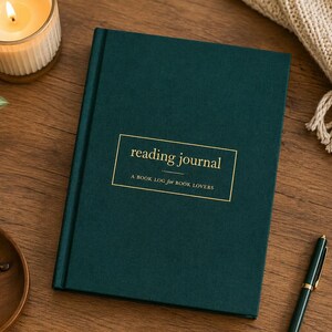 May include: A dark green hardcover reading journal with gold foil text on the front cover that reads reading journal A BOOK LOG for BOOK LOVERS. The journal is rectangular with a clean, solid-colored fabric texture and a gold-lined rectangular border.