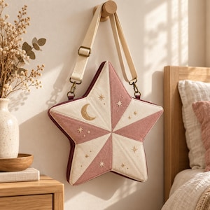 Peut inclure: Un sac bandoulière en forme d'étoile composé de panneaux alternés rose poudré et crème. Le sac est orné de motifs célestes dorés brodés, incluant un croissant de lune et diverses petites étoiles, et possède une bandoulière beige réglable.