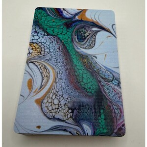 May include: A rectangular coaster with an abstract design. The artwork features a blend of colors, including teal, purple, gold, and white, creating a fluid, marbled effect. The coaster's edges are rounded.