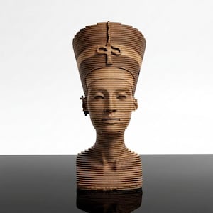 May include: A wooden sculpture of a bust of a woman with a tall headdress. The sculpture is made of layered wood, creating a striped effect. The bust is set against a white background and a black reflective surface.