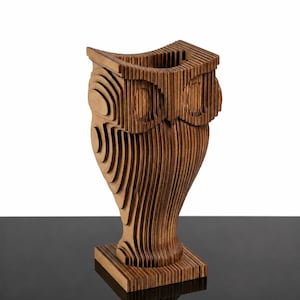 Puede incluir: Una escultura de búho de madera marrón construida con capas de madera contrachapada cortadas con láser y apiladas. El objeto presenta una abertura superior hueca, una base cuadrada y una textura estratificada que define la cabeza y el cuerpo.