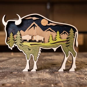 Peut inclure: Sculpture en bois en forme de bison avec un design en couches. L'œuvre d'art représente un paysage avec des montagnes, des arbres et deux bisons. Les couleurs incluent des tons de bois naturels, du vert et du bleu, créant une esthétique rustique.