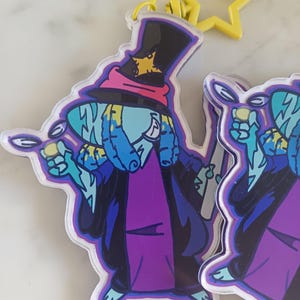 May include: An acrylic keychain featuring a cartoon character wearing a tall black top hat with a yellow star patch, a pink band, and a purple robe. The character has light blue skin, blue hair, and holds a magnifying glass. A yellow star-shaped clasp is attached.