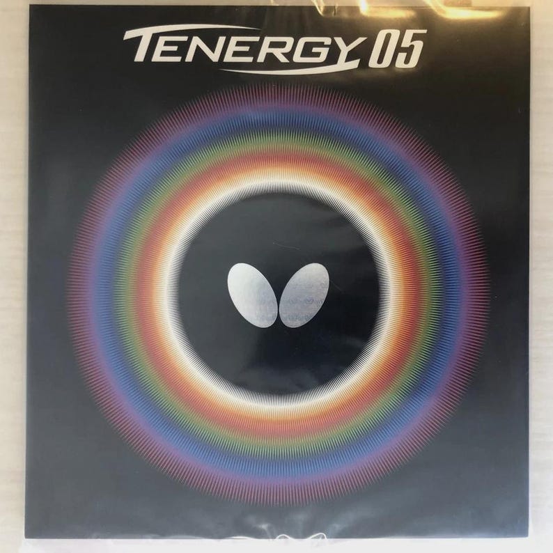 Op de afbeelding: Een vierkante zwarte productverpakking met de witte tekst TENERGY 05 bovenaan. Het midden toont een wit vlinderlogo omringd door een levendig, cirkelvormig regenboogkleurig gradi&euml;ntpatroon dat naar buiten straalt tegen de zwarte achtergrond.