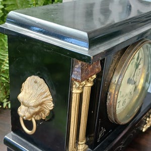 May include: A black mantel clock featuring gold-toned lion head side handles with rings, fluted columns, and ornate feet. The clock face is circular with a glass cover, and the black casing is decorated with thin white line accents and patterns.