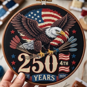 May include: A circular wooden embroidery hoop containing a cross-stitch design on black fabric. It features a bald eagle with spread wings, a United States flag, arrows, and the text 250 4TH JULY YEARS 2026 in red, white, blue, and gold.