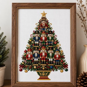 May include: A framed cross-stitch embroidery depicting a Christmas tree composed of multiple nutcracker figures in red, blue, green, and gold uniforms. The tree is decorated with small ornaments and topped with a gold star, all set in a dark wood frame.