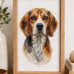 May include: A cross-stitch embroidery of a beagle dog head portrait featuring brown, black, and white thread colours on a white Aida fabric background, enclosed within a rectangular light-toned wooden frame with a visible grain texture.