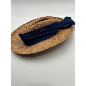 May include: A tan woven straw hat featuring a wide brim and a rounded crown. A dark blue fabric band wraps around the base of the crown, tied in a knot on the side with frayed edges. A small brown Nike swoosh logo is embroidered on the front band.