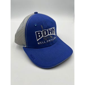 May include: A royal blue baseball cap with a grey mesh back and a flat brim. The front features white embroidered text that reads BOHN WELL DRILLING with a graphic of a drilling rig. The word MAX is embossed on the left side of the brim.