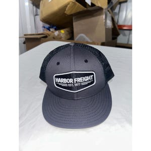 Może przedstawiać: Szara i czarna czapka typu trucker z napisem "HARBOR FREIGHT EST. 1977" na biało-czarnej naszywce. Czapka ma zakrzywiony daszek i siatkową tylną część.