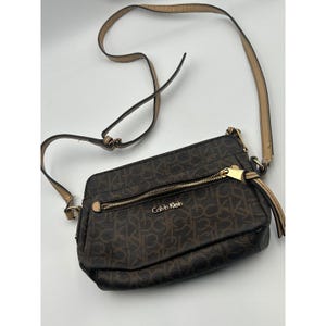 May include: A dark brown crossbody bag featuring the Calvin Klein logo pattern in a lighter brown. The bag includes a gold-tone zipper on the front pocket, a gold-tone Calvin Klein brand name, and a long, adjustable tan shoulder strap.