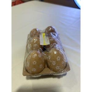 May include: A package of six decorative wooden eggs. Each egg is light brown and adorned with a pattern of small white flowers. The eggs are packaged in a cardboard container and wrapped in clear plastic.