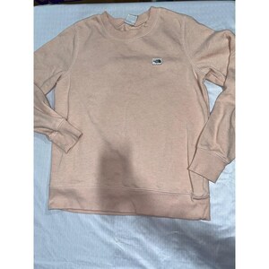 May include: A light peach-colored sweatshirt with a crew neck and long sleeves. A small, rectangular patch with a mountain graphic is on the upper left chest. The sweatshirt has ribbed cuffs and hem.