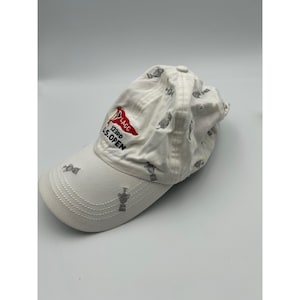 May include: A white baseball cap featuring an embroidered red flag with the text LACC and 123RD U.S. OPEN in black. The cap is decorated with a repeating pattern of small, grey embroidered trophy icons across the crown and the curved brim.