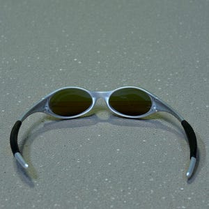 May include: Silver-framed sunglasses with oval lenses and a sleek, modern design. The lenses have a brown tint, and the temples feature black rubber grips for comfort. These sunglasses are a stylish accessory for outdoor activities.