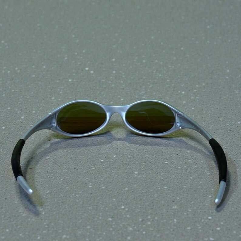 May include: Silver-framed sunglasses with oval lenses and a sleek, modern design. The lenses have a brown tint, and the temples feature black rubber grips for comfort. These sunglasses are a stylish accessory for outdoor activities.