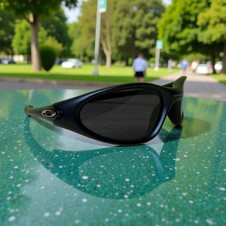 K&ouml;nnte beinhalten: Schwarze Sonnenbrille mit geschwungenem Rahmen und dunklen Gl&auml;sern. Die Sonnenbrille liegt auf einer gr&uuml;n gesprenkelten Oberfl&auml;che, mit einer Parkszene im Hintergrund. Sie hat ein kleines silbernes Emblem an der Seite.