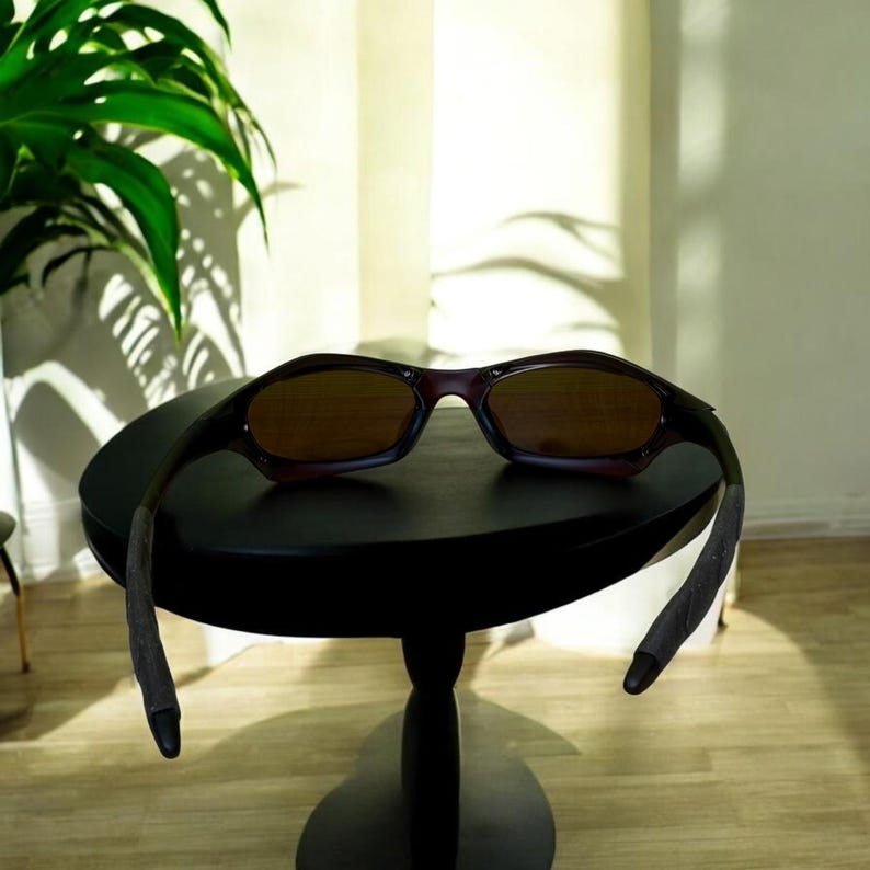 May include: Brown-tinted sunglasses with a dark frame rest on a small black table. The sunglasses have a curved design and are angled towards the viewer. The table is round and has a single pedestal base. The background includes a green plant and a white wall.