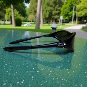 K&ouml;nnte beinhalten: Schwarze Sonnenbrille mit einem schlanken, modernen Design. Die Sonnenbrille hat dunkle Gl&auml;ser und einen schwarzen Rahmen mit silbernen Akzenten an den B&uuml;geln. Sie liegt auf einer gesprenkelten gr&uuml;nen Oberfl&auml;che.