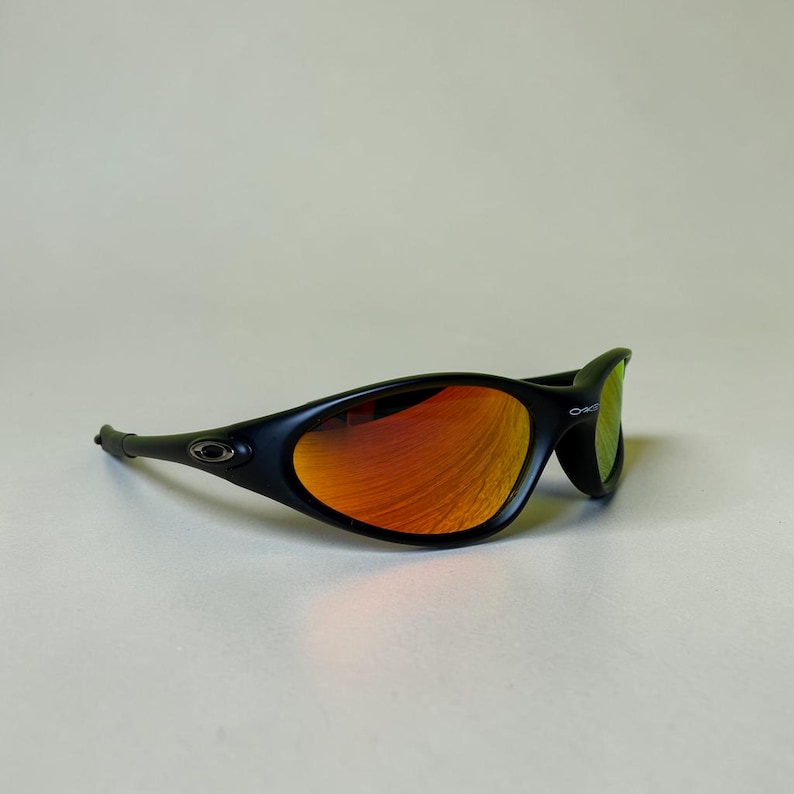 May include: Black sunglasses with orange mirrored lenses. The sunglasses have a sleek, curved design with a black frame. The lenses reflect a vibrant orange hue, and the Oakley logo is visible on the arm.