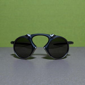 May include: Dark grey sunglasses with a distinctive design. The sunglasses feature a curved bridge and round lenses. The lenses are dark, and the frame has silver detailing.