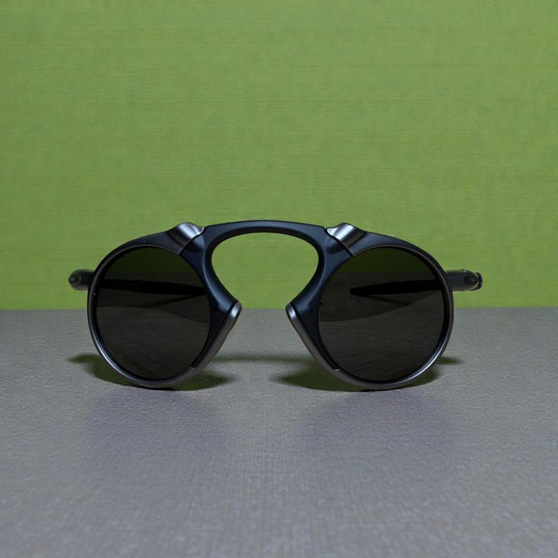 May include: Dark grey sunglasses with a distinctive design. The sunglasses feature a curved bridge and round lenses. The lenses are dark, and the frame has silver detailing.