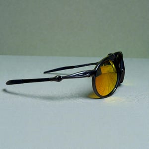 May include: Dark gray sunglasses with orange-tinted lenses. The sunglasses have a sleek, modern design with thin temples and a rounded frame. The lenses reflect a bright yellow color, and the frame appears to be made of metal.