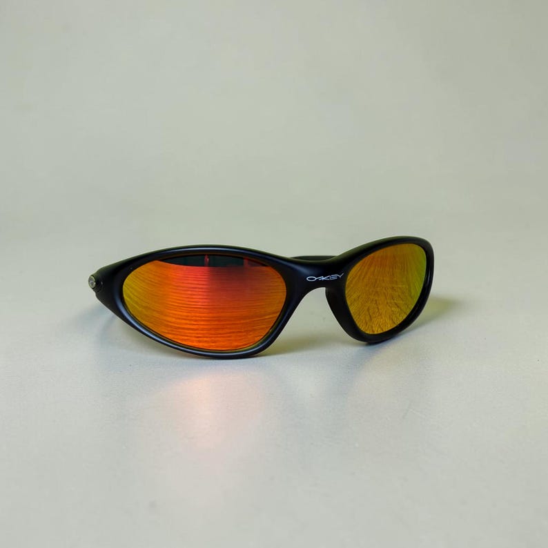 May include: Black sunglasses with orange-red mirrored lenses. The sunglasses have a curved, wraparound design and the brand name "Oakley" is visible on the frame. The lenses reflect a vibrant orange hue.