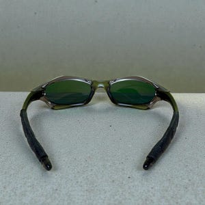 May include: Green-tinted sunglasses with a dark olive frame and black ear pieces. The lenses are a reflective green, and the frame has a sleek, modern design. The ear pieces are made of a textured, rubber-like material.