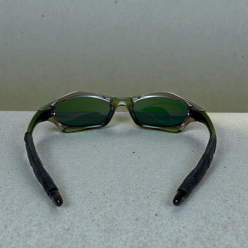 May include: Green-tinted sunglasses with a dark olive frame and black ear pieces. The lenses are a reflective green, and the frame has a sleek, modern design. The ear pieces are made of a textured, rubber-like material.