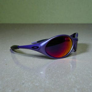 May include: Purple sunglasses with a sleek, oval frame and mirrored lenses that reflect shades of red and orange. The arms are a matching purple, with a black ear piece. The sunglasses are set against a neutral background.