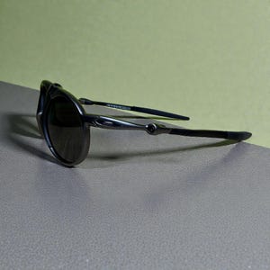 May include: Dark grey sunglasses with a curved frame and dark lenses. The temples are thin and black, with a silver accent near the hinge. The sunglasses are angled, showing the sleek design and the brand name on the temple.
