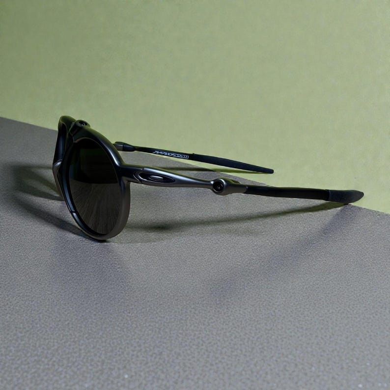 May include: Dark grey sunglasses with a curved frame and dark lenses. The temples are thin and black, with a silver accent near the hinge. The sunglasses are angled, showing the sleek design and the brand name on the temple.