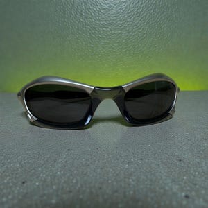 May include: Sleek, modern sunglasses with a silver frame and dark lenses. The glasses have a curved design and a subtle, metallic finish. The lenses are dark, providing shade and protection from the sun. The overall aesthetic is sporty and stylish.