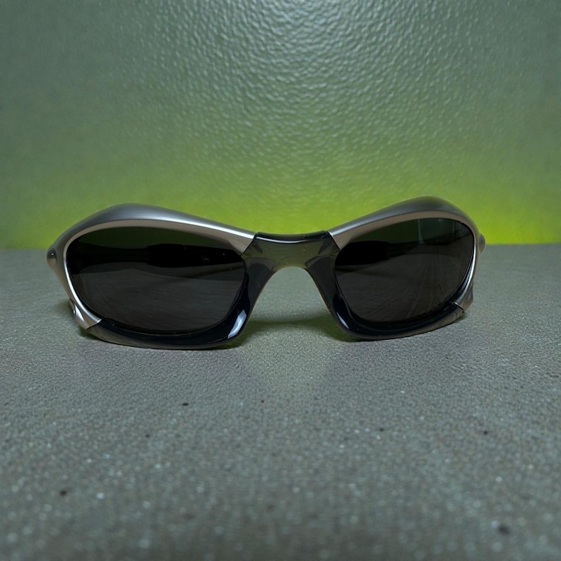 May include: Sleek, modern sunglasses with a silver frame and dark lenses. The glasses have a curved design and a subtle, metallic finish. The lenses are dark, providing shade and protection from the sun. The overall aesthetic is sporty and stylish.